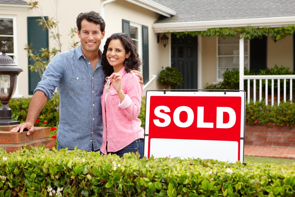 Hispanic couple outside home with sold sign.